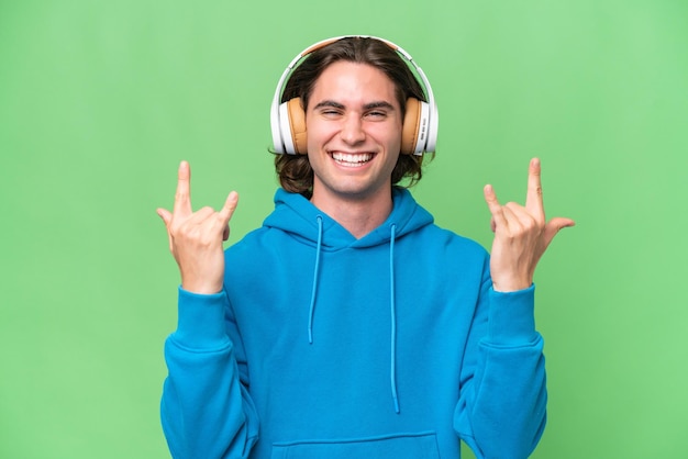 Young handsome man isolated on green chroma background listening music making rock gesture