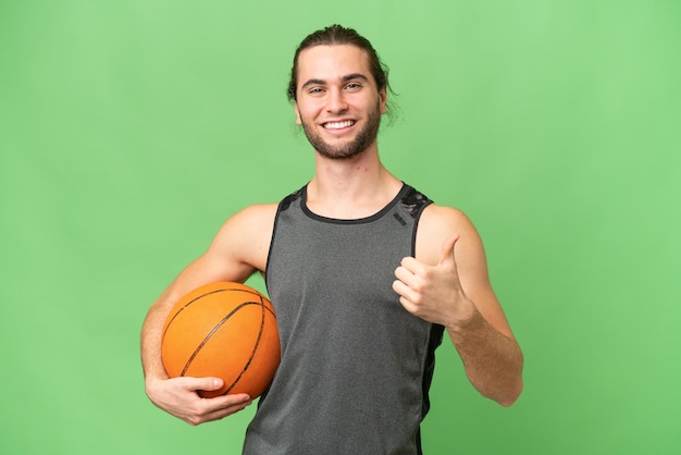 Young handsome man isolated on green chroma background playing basketball and with thumb up