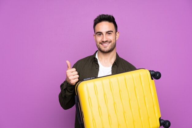 Young handsome man over isolated purple wall in vacation with travel suitcase and with thumb up