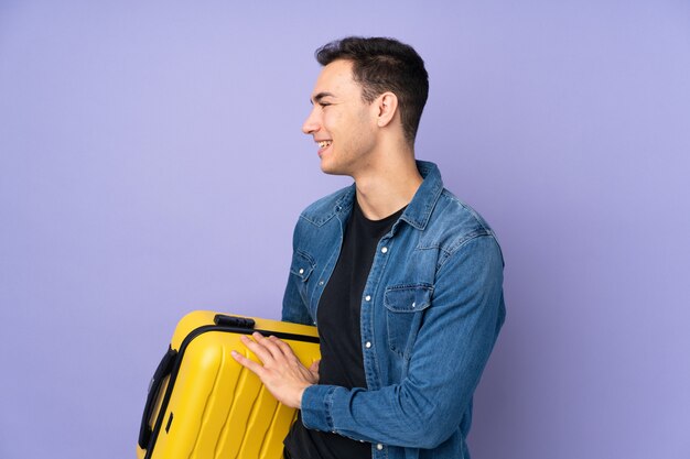 Young handsome man isolated on purple wall in vacation with travel suitcase