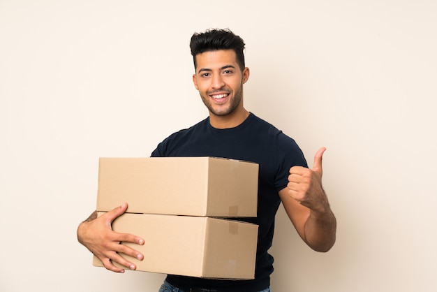 Young handsome man over isolated wall holding a box to move it to another site with thumb up
