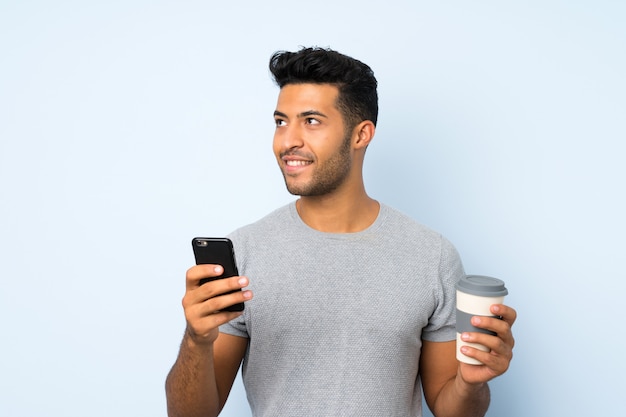 Young handsome man over isolated wall holding coffee to take away and a mobile while thinking something