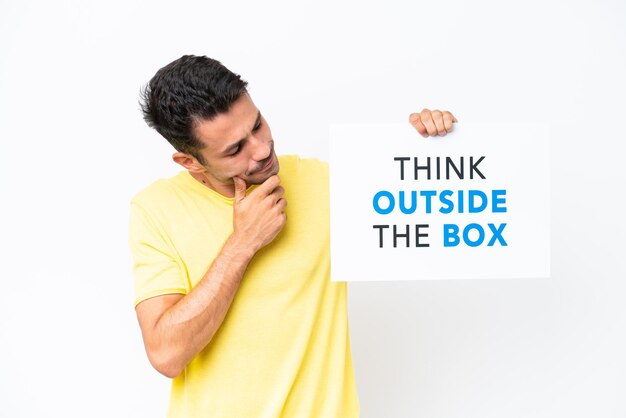 외진 흰색 배경 위에 있는 젊은 잘생긴 남자는 Think Outside The Box라는 텍스트가 있는 현수막을 들고 생각하고 있습니다.