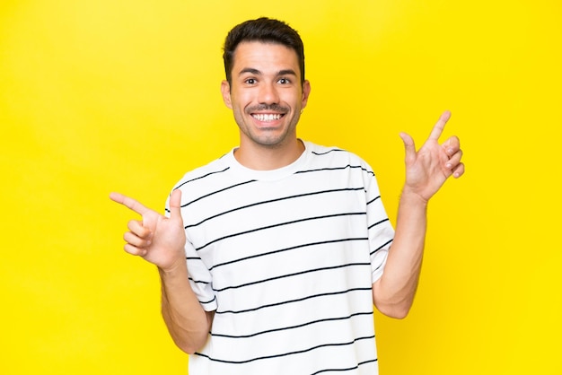 孤立した黄色の背景上の若いハンサムな男は、側面と幸せに指を指しています