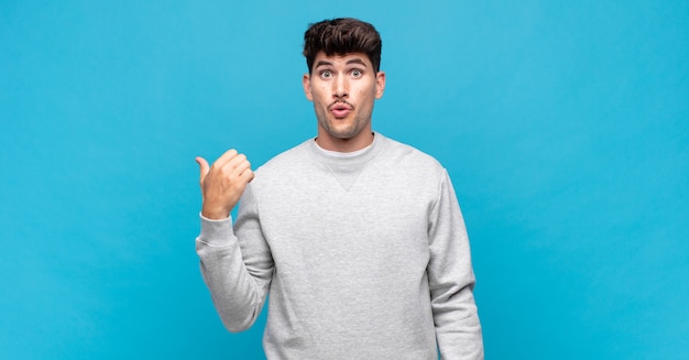 Young handsome man looking astonished in disbelief, pointing at object on the side