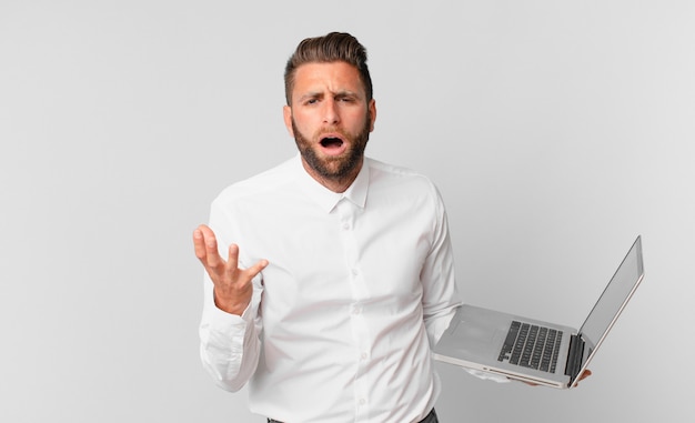 Young handsome man looking desperate, frustrated and stressed and holding a laptop