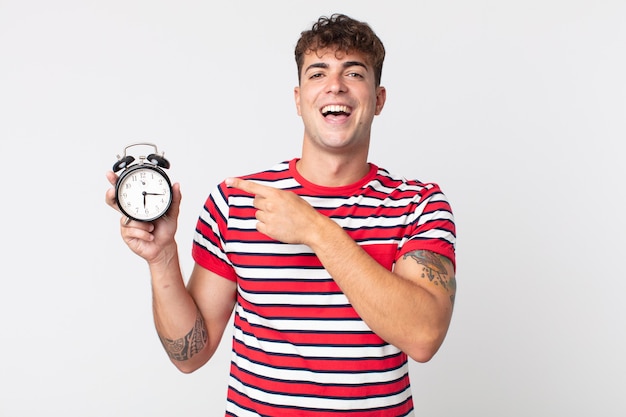 Young handsome man looking excited and surprised pointing to the side and holding an alarm clock