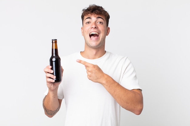 Young handsome man looking excited and surprised pointing to the side and holding a beer bottle