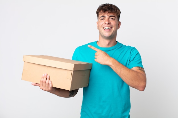 Young handsome man looking excited and surprised pointing to the side and holding a cardboard box