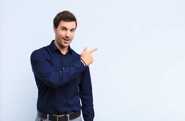 Young handsome man looking excited and surprised pointing to the side and upwards to copy space against blue wall