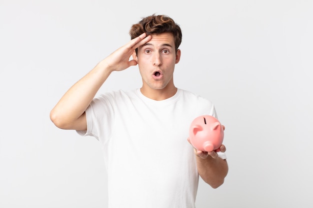 Young handsome man looking happy, astonished and surprised and holding a piggy bank