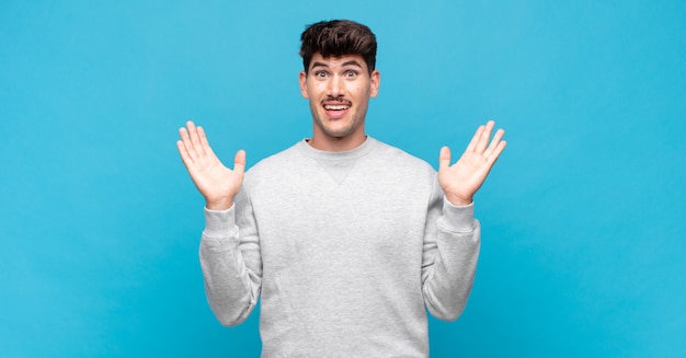 Young handsome man looking happy and excited, shocked with an unexpected surprise with both hands open next to face