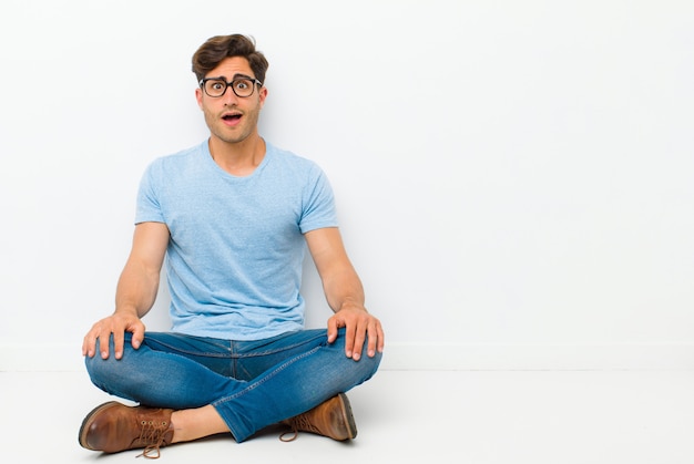 Young handsome man looking happy and pleasantly surprised, excited with a fascinated and shocked expression sitting on the floor