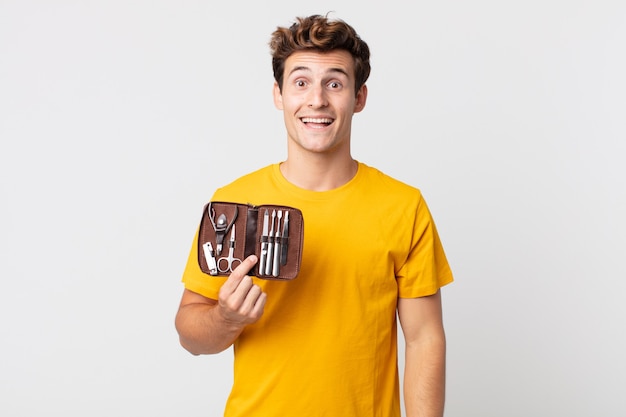 Young handsome man looking happy and pleasantly surprised and holding a nails tools case