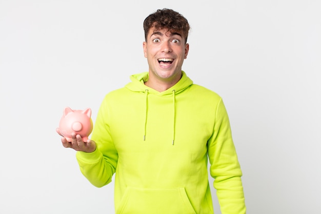 Young handsome man looking happy and pleasantly surprised and holding a piggy bank