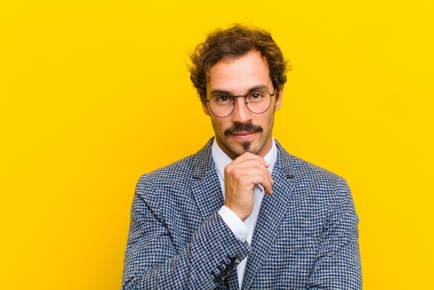 Young handsome man looking happy and smiling with hand on chin, wondering or asking a question, comparing options against orange 