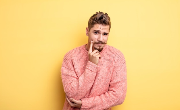 Young handsome man looking happy and smiling with hand on chin wondering or asking a question comparing options strange moustache concept