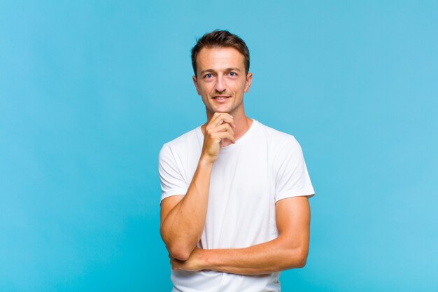 Young handsome man looking happy and smiling with hand on chin, wondering or asking a question, comparing options