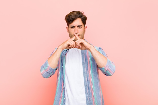 Young handsome man looking serious and displeased with both fingers crossed up front in rejection, asking for silence against pink background