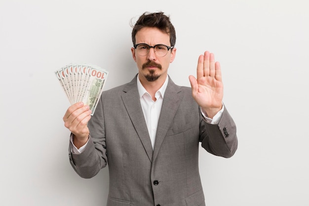 Young handsome man looking serious showing open palm making stop gesture businessman and banknotes