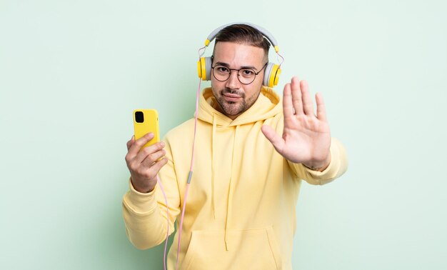 Young handsome man looking serious showing open palm making stop gesture. headphones and smartphone concept