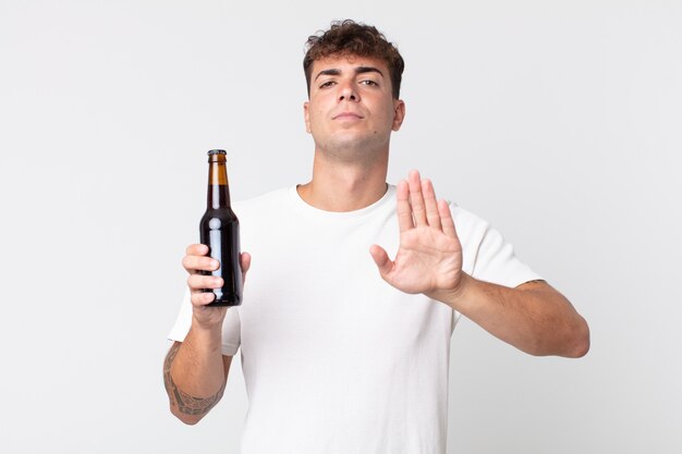 真剣に見える若いハンサムな男は、開いた手のひらを停止ジェスチャーをし、ビール瓶を持っていることを示しています