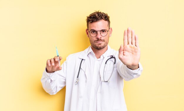 Giovane uomo bello che sembra serio mostrando palmo aperto che fa gesto di arresto. concetto di siringa del medico