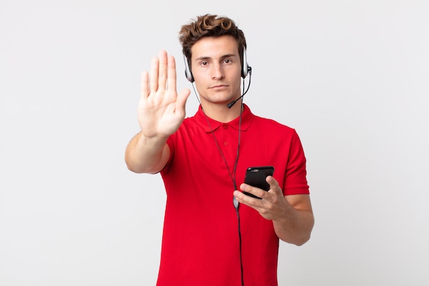 Young handsome man looking serious showing open palm making stop gesture with a smartphone and headset