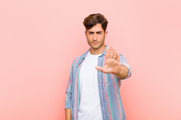 Young handsome man looking serious, stern, displeased and angry showing open palm making stop gesture against pink wall