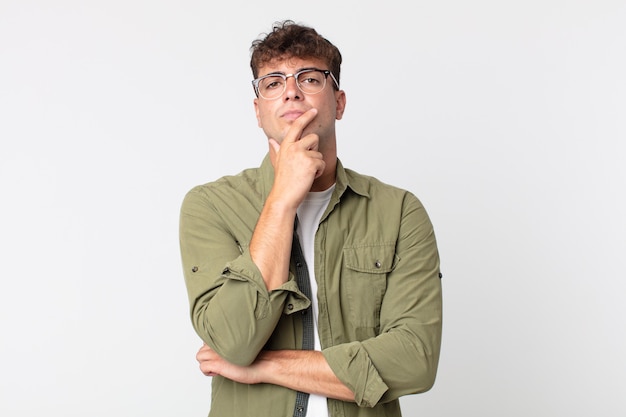 Young handsome man looking serious, thoughtful and distrustful, with one arm crossed and hand on chin, weighting options