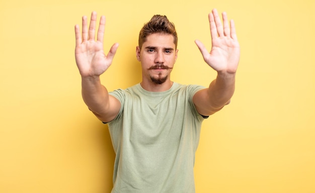 Young handsome man looking serious, unhappy, angry and displeased forbidding entry or saying stop with both open palms. strange moustache concept