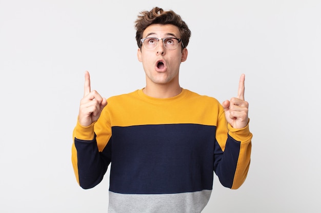 Young handsome man looking shocked, amazed and open mouthed, pointing upwards with both hands to copy space