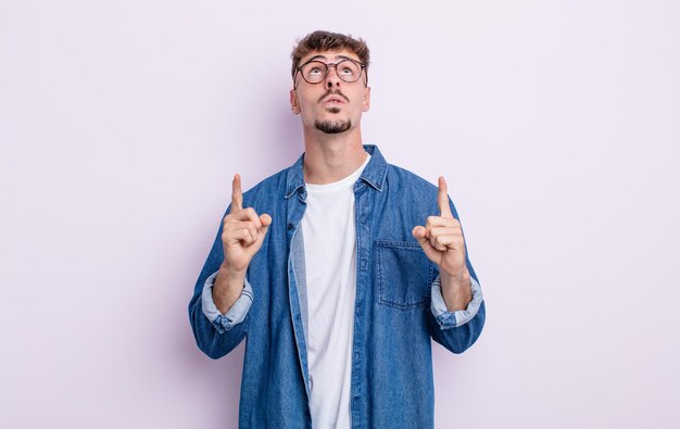 Young handsome man looking shocked, amazed and open mouthed, pointing upwards with both hands to copy space