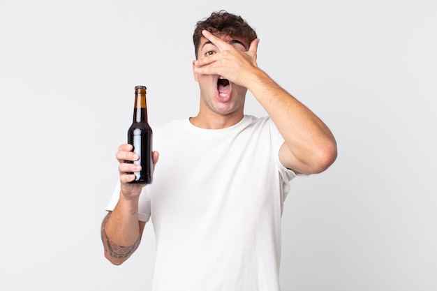 Young handsome man looking shocked, scared or terrified, covering face with hand and holding a beer bottle