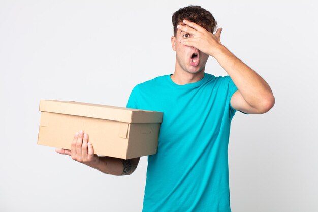 Young handsome man looking shocked scared or terrified covering face with hand and holding a cardboard box