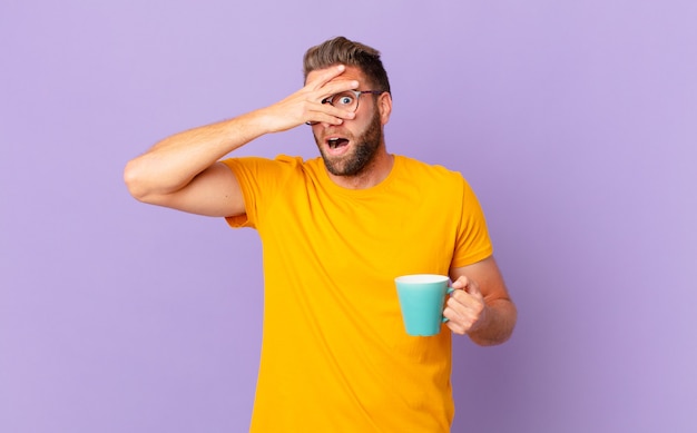 Young handsome man looking shocked, scared or terrified, covering face with hand. and holding a coffee mug