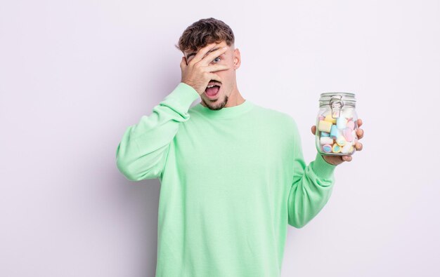 Young handsome man looking shocked, scared or terrified, covering face with hand. jelly candies concept