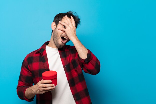 Young handsome man looking shocked, scared or terrified, covering face with hand and peeking between fingers