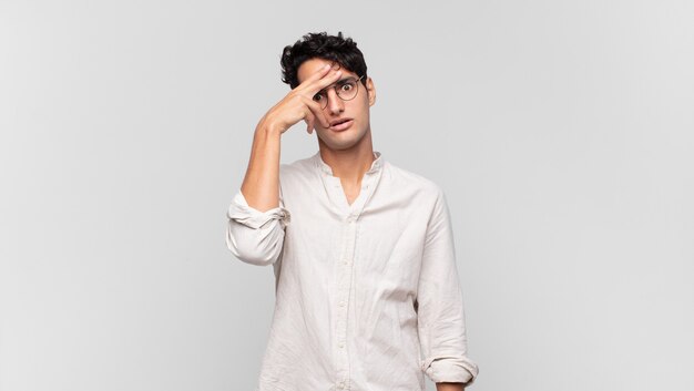 Young handsome man looking shocked, scared or terrified, covering face with hand and peeking between fingers