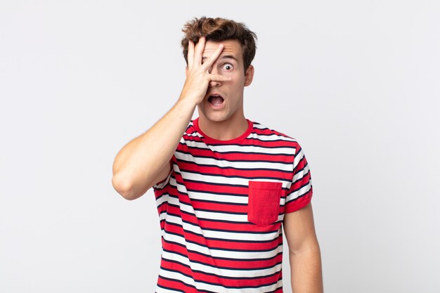 young handsome man looking shocked, scared or terrified, covering face with hand and peeking between fingers