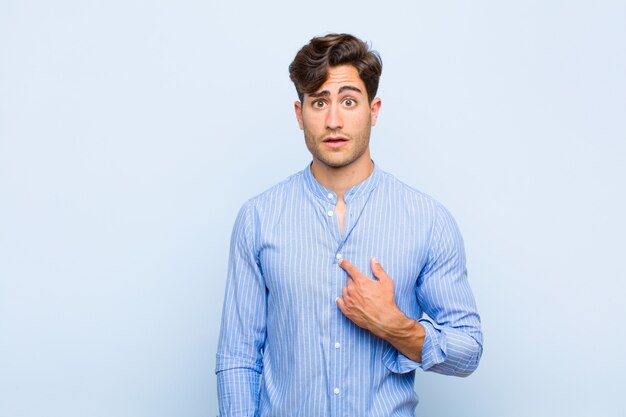 Young handsome man looking shocked and surprised with mouth wide open, pointing to self against blue background