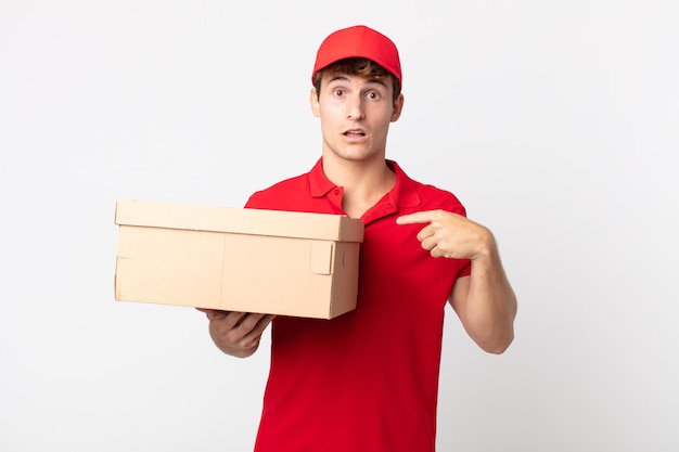 Young handsome man looking shocked and surprised with mouth wide open, pointing to self delivery package service concept.