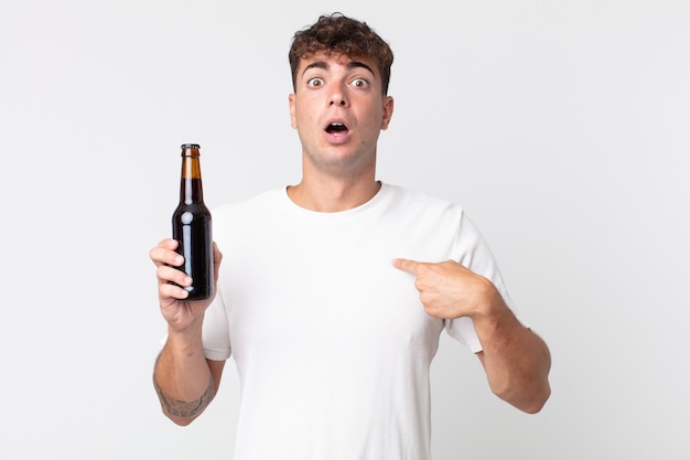 Young handsome man looking shocked and surprised with mouth wide open, pointing to self and holding a beer bottle