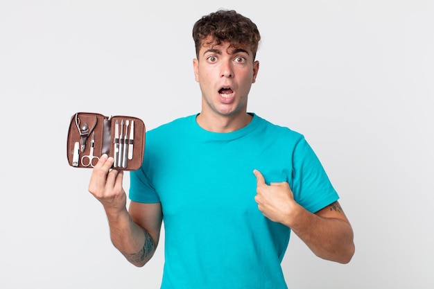 Young handsome man looking shocked and surprised with mouth wide open, pointing to self and holding a nails tools case