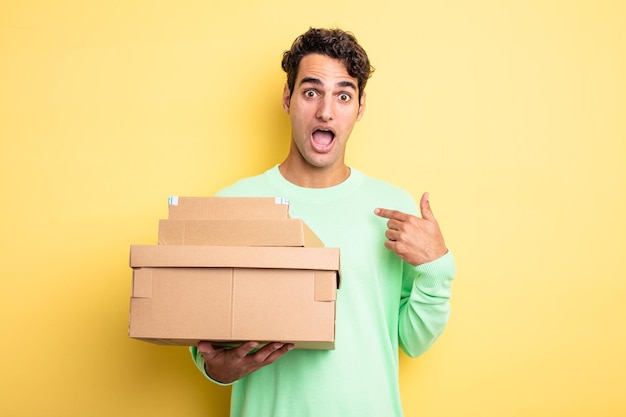 Young handsome man looking shocked and surprised with mouth wide open pointing to self package boxes