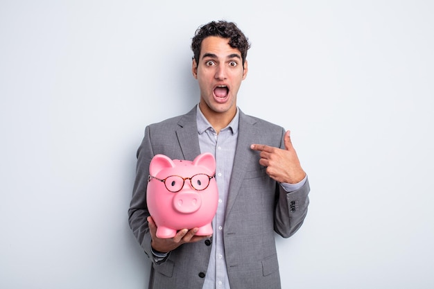 Young handsome man looking shocked and surprised with mouth wide open pointing to self piggy bank concept