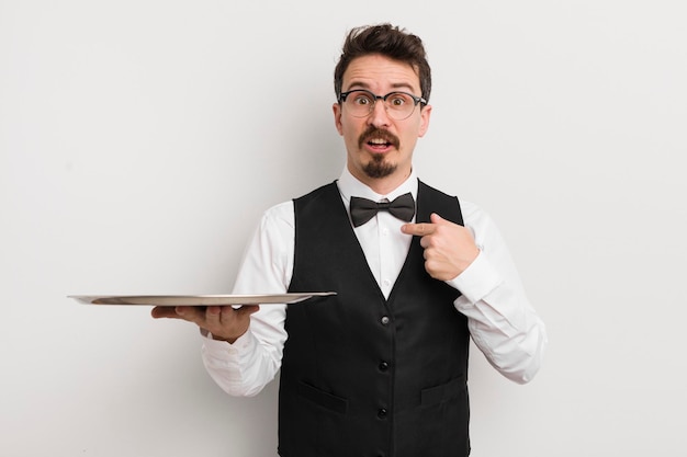 Young handsome man looking shocked and surprised with mouth wide open pointing to self waiter and tray concept