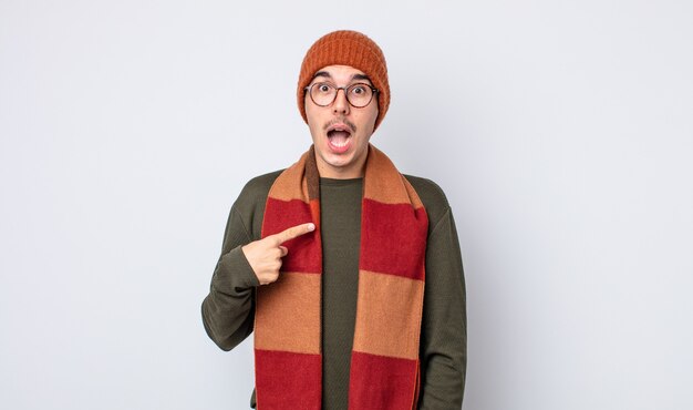 young handsome man looking shocked and surprised with mouth wide open, pointing to self. winter clothes concept