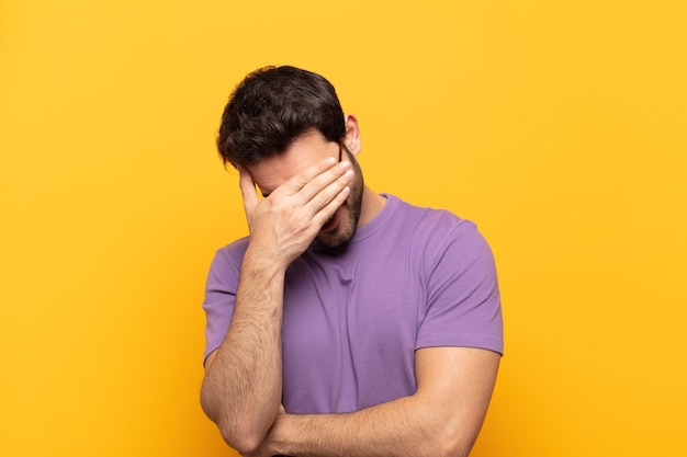 Giovane uomo bello che sembra stressato, vergognoso o turbato, con un mal di testa, che copre il viso con la mano