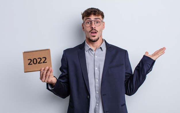 Young handsome man looking surprised and shocked, with jaw dropped holding an object. calendar concept